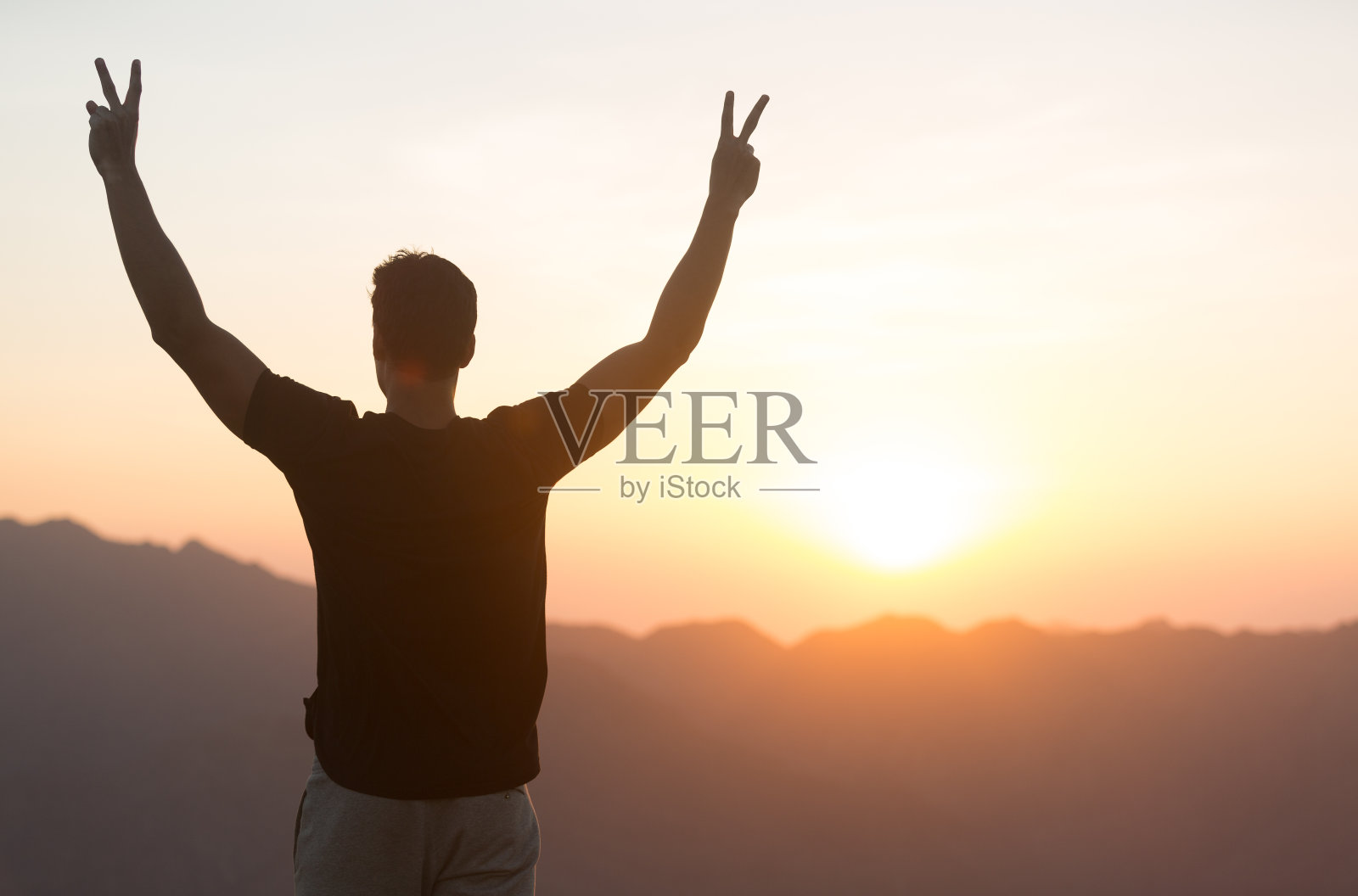 日落时分，一个快乐的男人在山顶上举起手臂庆祝。照片摄影图片