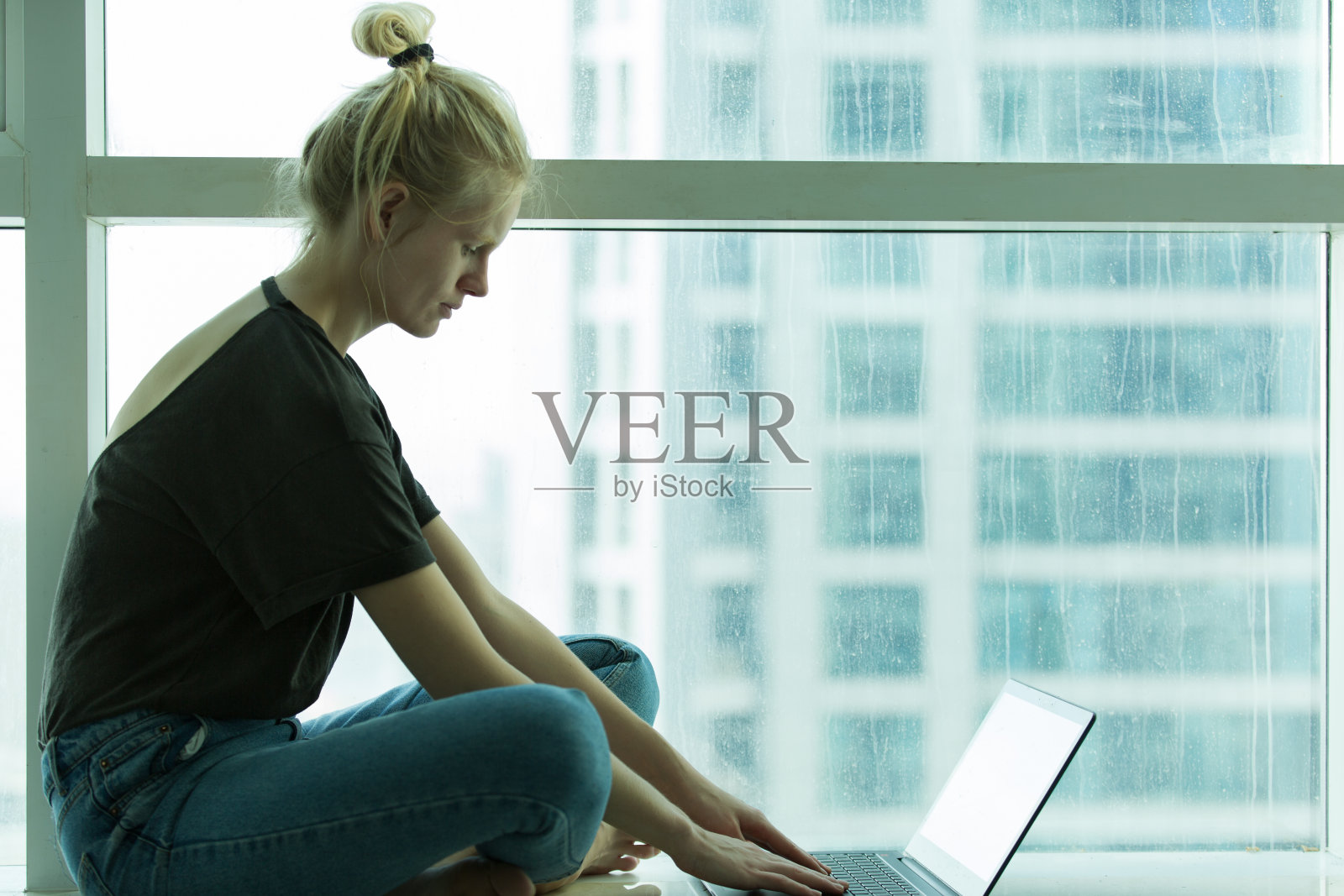 放松的年轻女子打字笔记本电脑坐在旁边的公寓窗户。照片摄影图片