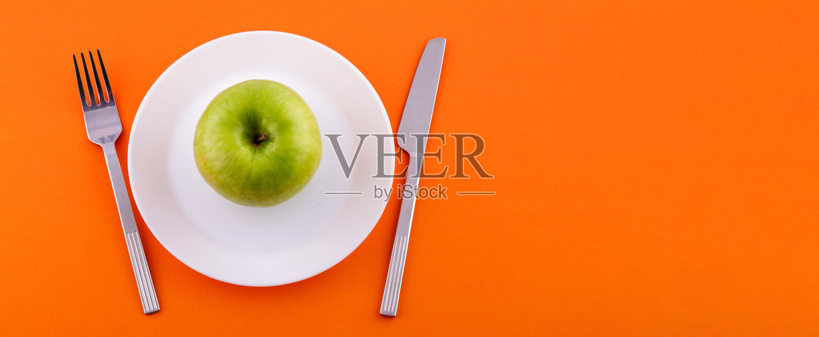 一个白色的盘子上放着一个青苹果，一把带叉的刀放在橙色的背景上，从上面看。饮食与健康食品理念照片摄影图片