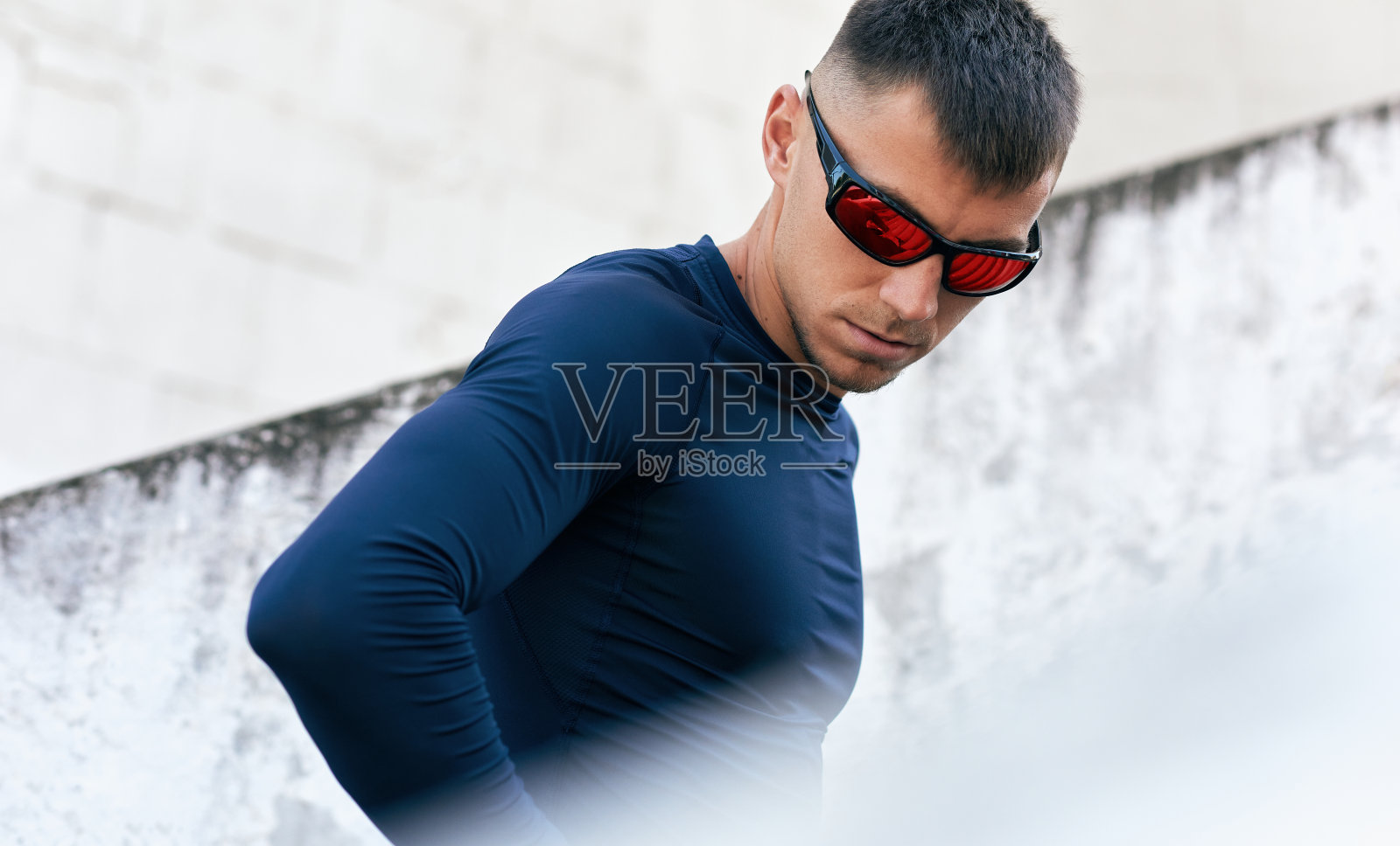 健康的运动员，穿着蓝色运动服，戴着太阳镜，看着别处，站在城市的街道上。运动员胳膊搂在腰上在街上休息。照片摄影图片