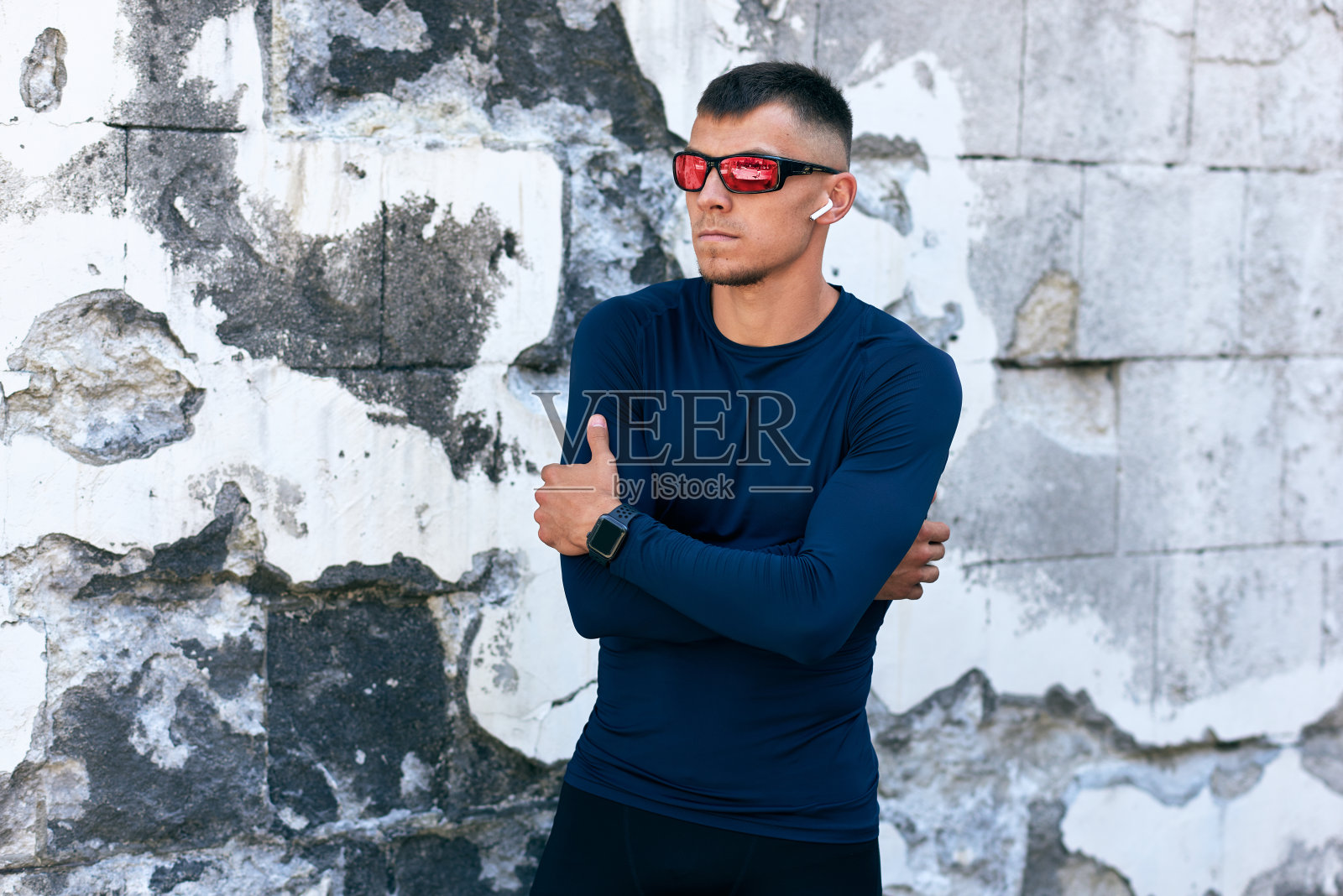 户外肖像的fit年轻男子在蓝色运动服和红色太阳镜看向别处和站在城市的混凝土墙。运动员交叉双臂休息。照片摄影图片