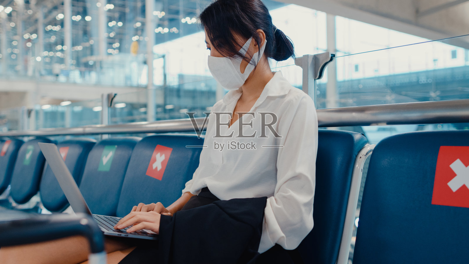 亚洲商务女旅行者戴口罩坐在长凳上用笔记本电脑在候机大厅等待航班。照片摄影图片