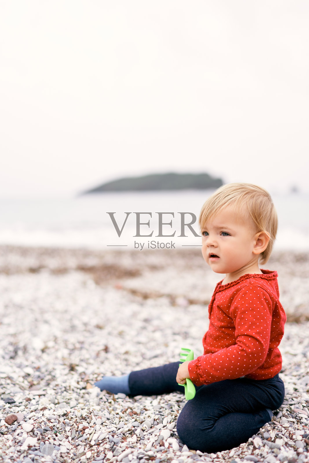 一个小孩坐在卵石滩上，手里拿着一个玩具耙子照片摄影图片