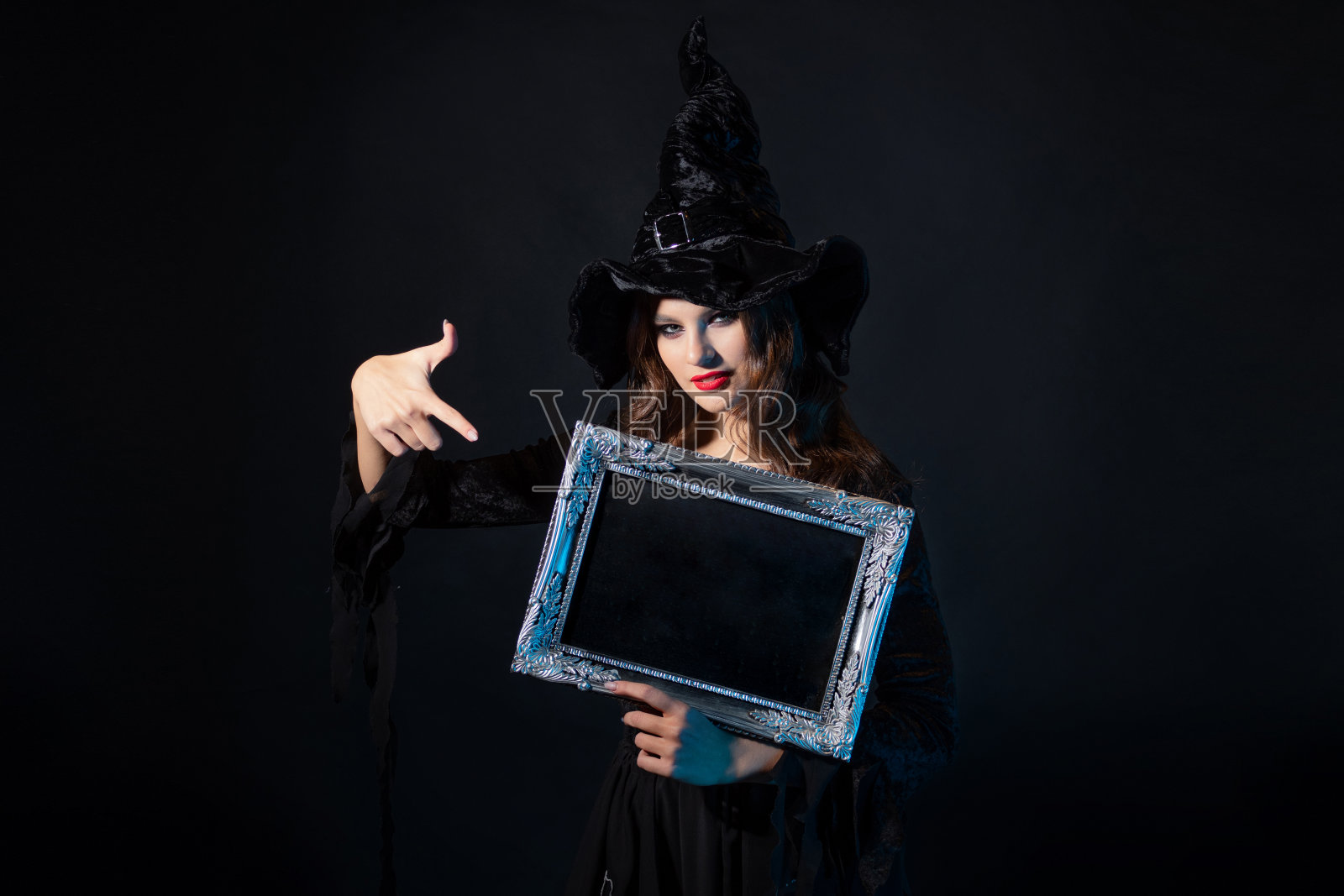 一个年轻美丽的女巫戴着尖顶的帽子，手里拿着一个银色的镜框，镜框上有黑色的字体，照片摄影图片