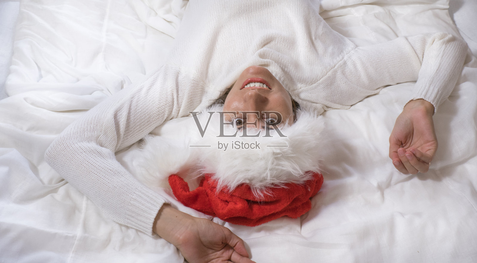 一个美丽的黑发女人戴着圣诞老人的帽子躺在白色的床上，看着镜头微笑。圣诞快乐。照片摄影图片