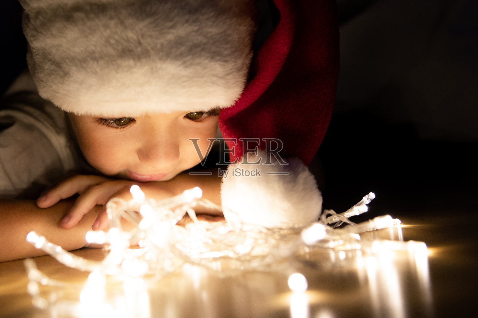 一个戴着圣诞帽的孩子在黑暗中趴在地板上，看着圣诞彩灯。孩子看着灯泡，想着礼物。抱臂下巴照片摄影图片