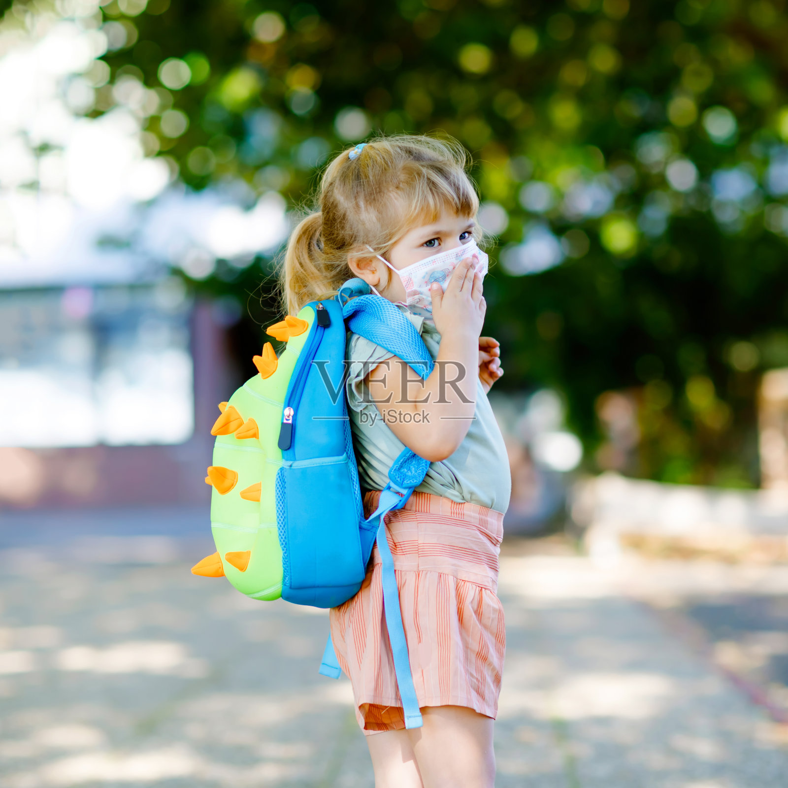 第一天上学的小女孩戴着抗冠状病毒的医用口罩。健康漂亮的宝宝步行去幼儿园、学前班和幼儿园。背背包的快乐孩子照片摄影图片
