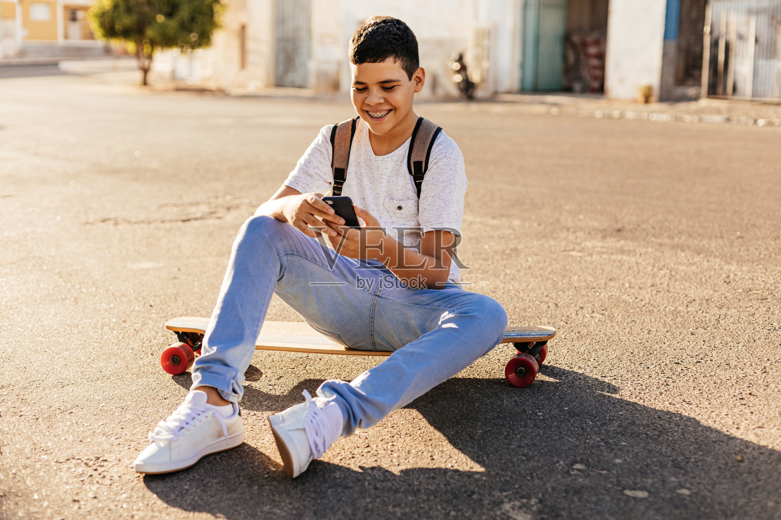 十几岁的男孩坐在街上的滑板上玩智能手机照片摄影图片