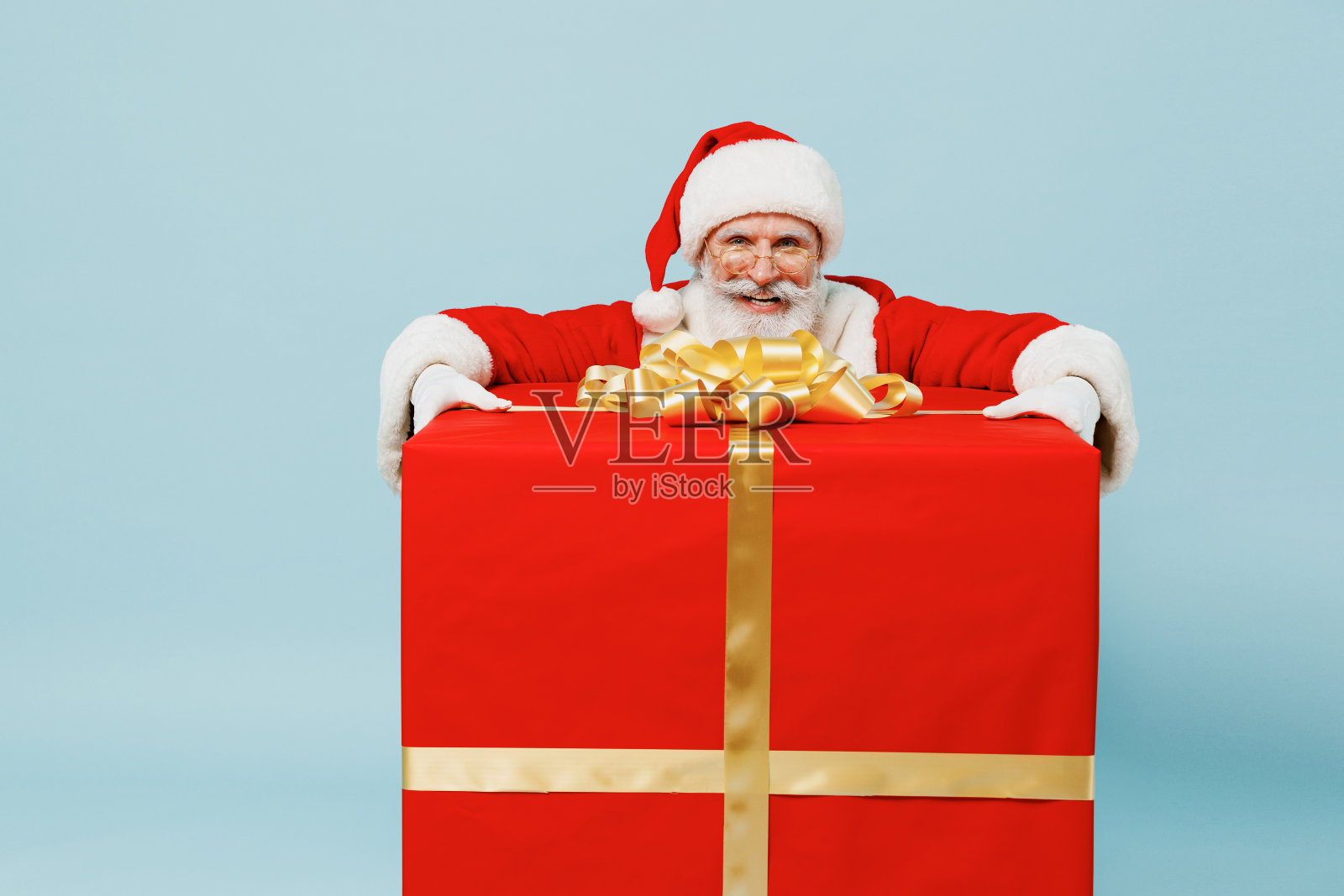 全身上下的圣诞老人50多岁，戴着圣诞帽，穿着红色西装，站在大礼物盒旁边，孤立在朴素的蓝色背景下，工作室新年快乐，2022年庆祝，圣诞快乐的节日概念照片摄影图片