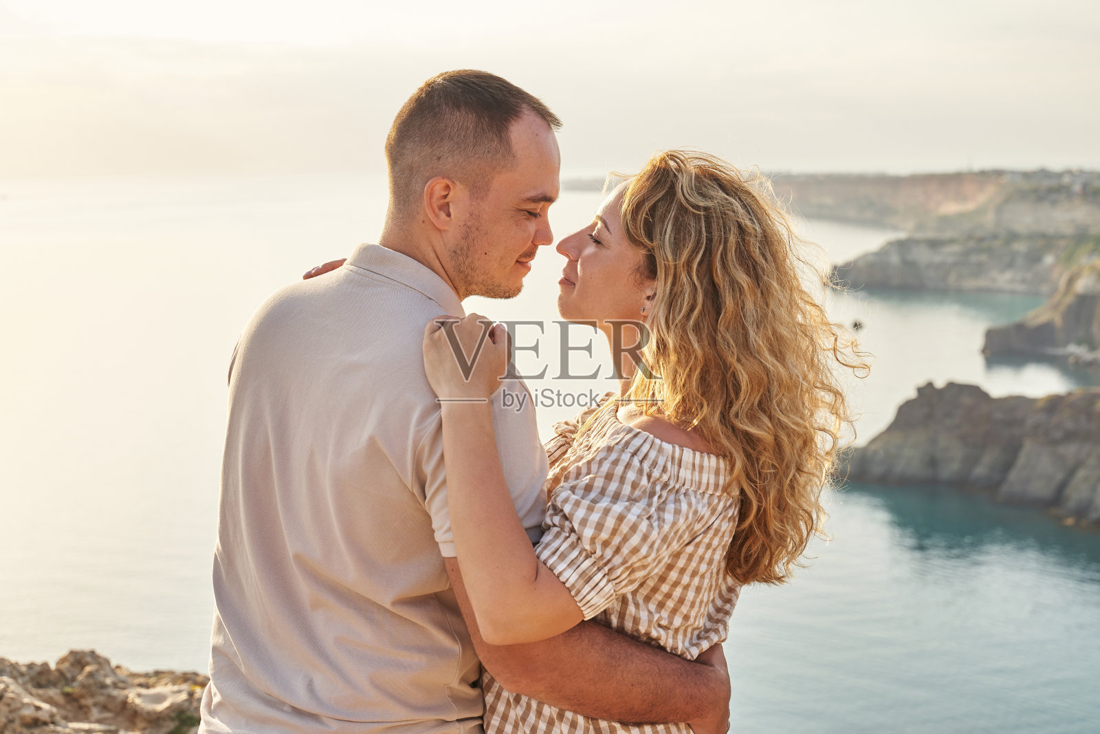 一对美丽的年轻夫妇站在岩石海岸，在夕阳的光线中面对面，试图亲吻。他们拥抱，女人怀孕了。爱、幸福照片摄影图片