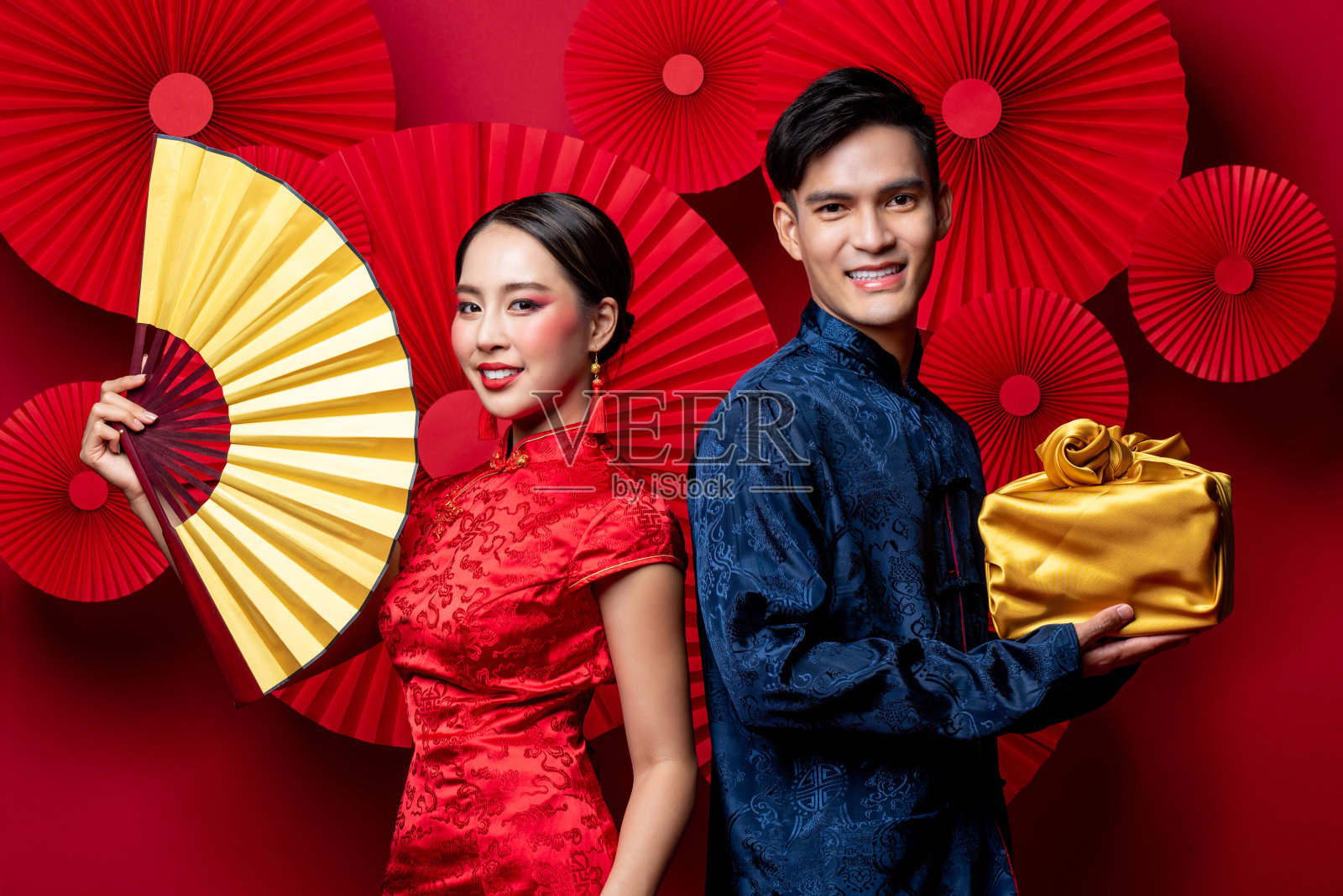身着传统服饰的亚洲夫妇面带微笑，以东方红色为背景装饰，以扇子和金色包装作为春节礼物照片摄影图片
