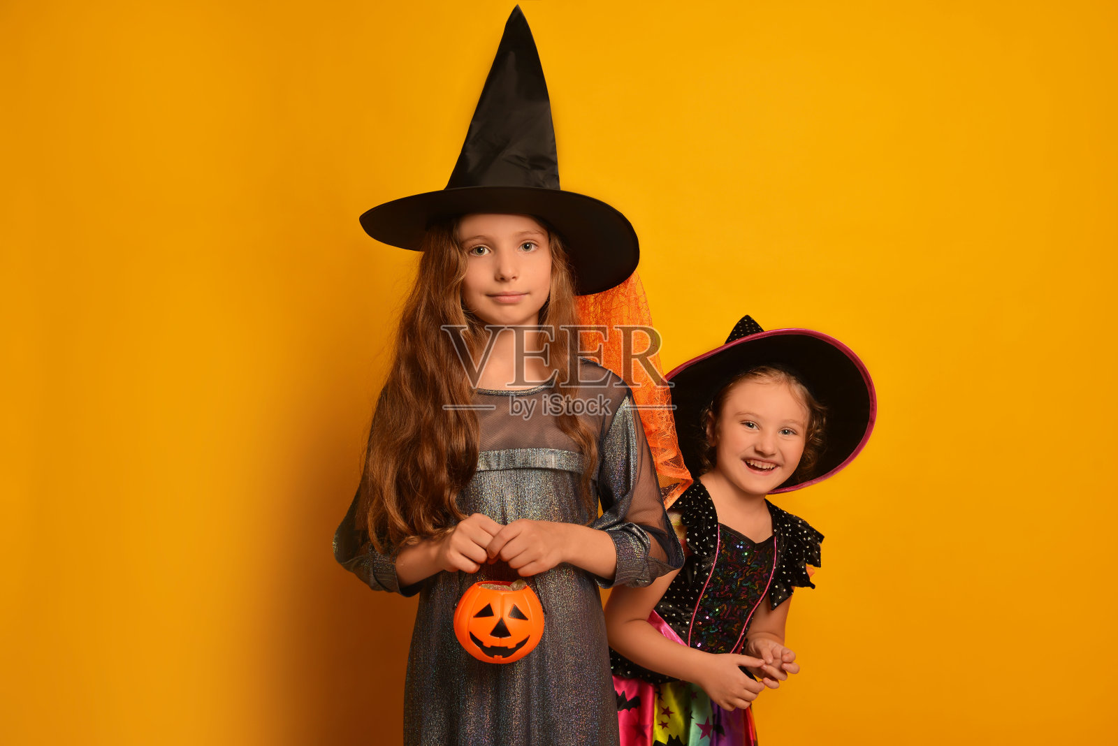 两个可爱的女孩姐妹打扮成万圣节的女巫或巫师在黄色的背景。

穿着狂欢节服装的孩子们拿着南瓜糖篮子。照片摄影图片